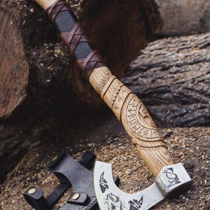 May include: A handcrafted axe with a wooden handle, wrapped in brown and black leather. The axe head is silver with a detailed engraved design. A black leather sheath is included.