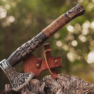 May include: A handcrafted axe with a silver blade featuring an intricate design. The wooden handle is dark brown with carved details and wrapped in tan leather. A brown leather sheath is attached.