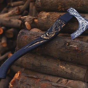 May include: A handcrafted axe with a dark blue handle and head. The axe head features intricate silver-colored designs. The handle is adorned with gold-colored carvings. The axe is resting on a pile of wooden logs.