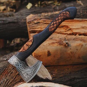 May include: A handcrafted axe with a black handle and a decorative, engraved blade. The handle features a carved wooden grip with a diamond pattern. The axe head has intricate silver designs. The axe is resting on a wooden log.