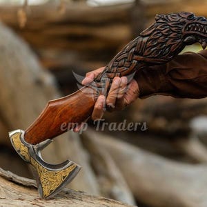 May include: A handcrafted axe with a wooden handle and a wolf head carving. The axe head is silver and gold with intricate designs. The handle is a rich brown color. The axe is held in a person's hand.