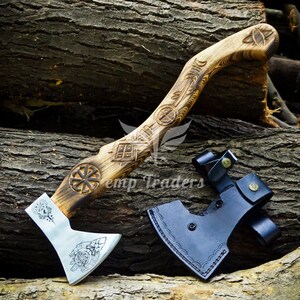 May include: Axe with a wooden handle featuring carved designs and a silver blade. The handle has intricate carvings, including circular and geometric patterns. A black leather sheath is included. The axe is displayed on a background of wooden logs.