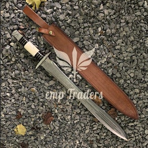 May include: A silver dagger with a bone and black handle, resting on a bed of gray gravel. The dagger is next to a brown leather sheath with a strap. The text "emp Traders" is visible.