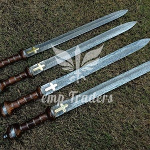 May include: Four swords with textured blades and wooden handles. Each sword features a gold-colored cross near the handle. The handles are a dark brown color with silver accents. The swords are displayed on a bed of grass.