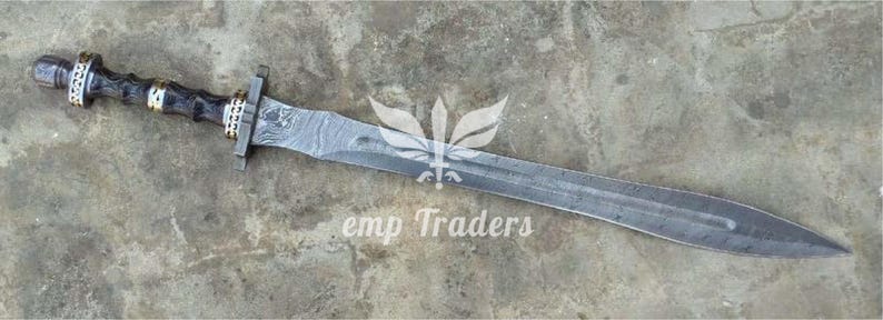 Puede incluir: Una espada detallada con una hoja estampada y un mango oscuro y texturizado. El mango presenta anillos decorativos con detalles dorados. El texto "emp Traders" es visible en la hoja.