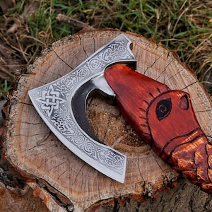 May include: A handcrafted axe with a silver blade featuring intricate Celtic knotwork and a curved cutting edge. The handle is carved from reddish-brown wood, shaped like a serpent's head. The axe rests on a weathered wooden stump, showcasing the details of the craftsmanship.