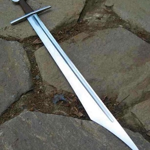 May include: A long, silver sword with a brown handle and a crossguard. The blade is sharp and pointed, and the sword is lying on a stone surface. The sword appears to be a historical or fantasy weapon.