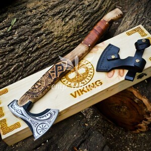May include: A Viking axe with a wooden handle and a silver blade, resting on a wooden box. The handle is wrapped in brown leather. The box has the word "VIKING" engraved on it, along with other Viking symbols. A black leather sheath is also included.