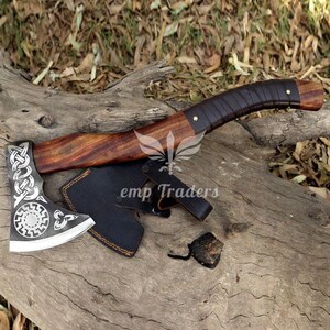 May include: Axe with a wooden handle wrapped in dark brown leather. The axe head is silver with an intricate design. A black leather sheath protects the blade. The axe rests on a weathered wooden surface.