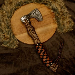 May include: A handcrafted axe with a patterned wooden handle and a checkered leather grip. The axe head is silver with etched designs. The axe is displayed on a wooden round surface with a fur-like background.