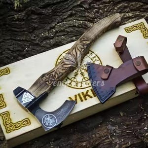 May include: A handcrafted Viking axe with a wooden handle featuring intricate carvings. The axe head is dark metal with etched designs. A brown leather sheath is included. The axe rests on a wooden box with Viking symbols.