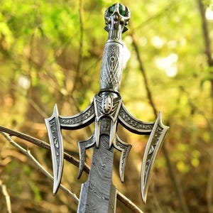 May include: A detailed close-up of a fantasy sword with an ornate silver handle and guard. The blade has a patterned design. The hilt features a green gem. The sword is set against a blurred green background of foliage.