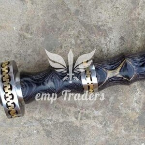 Puede incluir: Primer plano de una empu&ntilde;adura de espada y parte de la hoja. La empu&ntilde;adura presenta un mango de madera oscura estampada con detalles dorados y plateados. La hoja tiene un patr&oacute;n texturizado &uacute;nico. El texto "emp Traders" es visible.