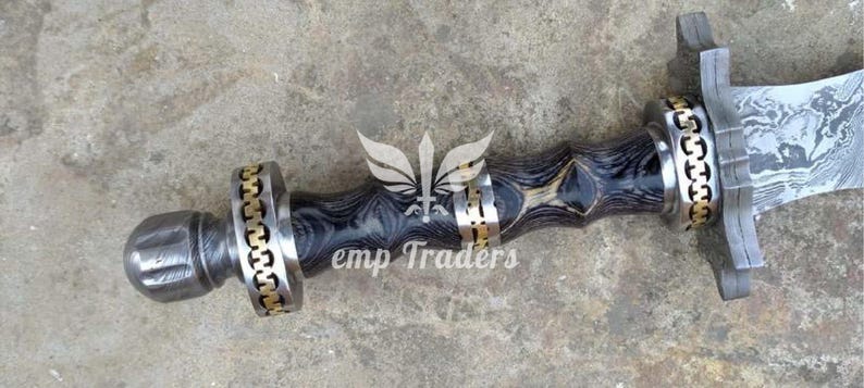 Puede incluir: Primer plano de una empu&ntilde;adura de espada y parte de la hoja. La empu&ntilde;adura presenta un mango de madera oscura estampada con detalles dorados y plateados. La hoja tiene un patr&oacute;n texturizado &uacute;nico. El texto "emp Traders" es visible.