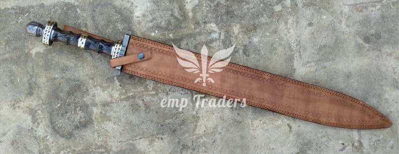 Puede incluir: Una espada romana gladius con una funda de cuero marr&oacute;n. La espada tiene un mango negro y plateado con detalles dorados. La funda est&aacute; grabada con un patr&oacute;n decorativo y el texto "emp Traders".