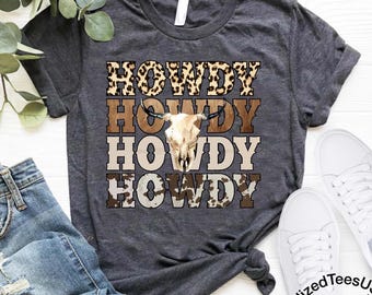 Könnte beinhalten: Dunkelgraues T-Shirt mit dem Wort "HOWDY" in Leopardenmuster, Braun, Beige und Kuhfellmuster. Ein Kuhschädel ist auf dem T-Shirt zentriert. Das T-Shirt wird mit blauen Jeans und weißen Turnschuhen kombiniert.