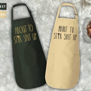 May include: Two black and beige aprons with the text "About to Stir Shit Up" printed on them. The aprons have adjustable neck straps and a pocket on the front.
