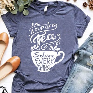 May include: A navy blue t-shirt with the text "A Cup of Tea Solves Everything" in white script. The image includes a pair of brown flats and a pair of blue jeans.