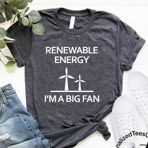 May include: Dark gray t-shirt with the text "RENEWABLE ENERGY I'M A BIG FAN" and a graphic of two white wind turbines. The shirt is paired with blue jeans and white sneakers, suggesting casual wear.