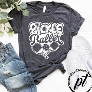 以下が含まれることがあります： 「Pickle Baller」の白い文字と、ピックルボールのラケットとボールのグラフィックがプリントされた濃いグレーのTシャツ。青いジーンズと白いスニーカーが写っています。Tシャツはカジュアルなスタイルです。