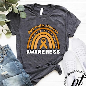 May include: Dark gray t-shirt with a rainbow design in shades of orange and the words "Appendix Cancer Awareness". The design includes a ribbon symbol. The shirt is paired with blue jeans and white sneakers.