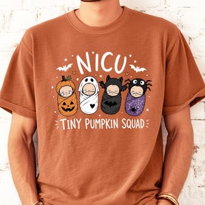 May include: A rust-colored t-shirt featuring a graphic of four swaddled babies in Halloween costumes, including a pumpkin, ghost, bat, and spider. The text reads NICU and TINY PUMPKIN SQUAD, accompanied by small stars, hearts, and bat silhouettes.