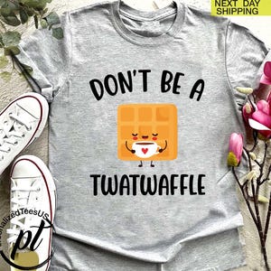 K&ouml;nnte beinhalten: Hellgraues T-Shirt mit dem schwarzen Schriftzug "DON'T BE A TWATWAFFLE". In der Mitte befindet sich eine Cartoon-Waffel, die eine Kaffeetasse h&auml;lt. Wei&szlig;e Turnschuhe sind links. "NEXT DAY SHIPPING" ist oben rechts.