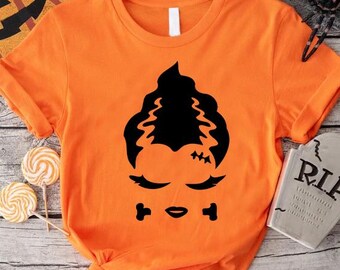 May include: Orange t-shirt with a black graphic of a Frankenstein's monster face. The design includes the monster's hair, eyes, mouth, and stitches. Halloween-themed decorations, including a pumpkin, spider, and tombstone, are in the background.