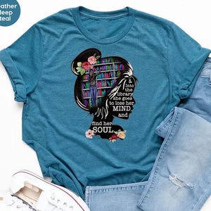 May include: Heather deep teal t-shirt with a graphic of a woman's silhouette with a bookshelf inside her head, flowers, and the text "& into the library she goes to lose her MIND and find her SOUL." Light blue jeans and white sneakers are also in the image.