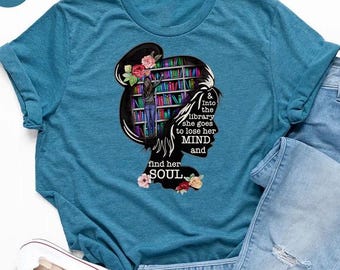 May include: Heather deep teal t-shirt with a graphic of a woman's silhouette with a bookshelf inside her head, flowers, and the text "& into the library she goes to lose her MIND and find her SOUL." Light blue jeans and white sneakers are also in the image.