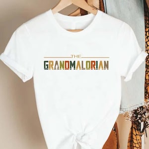 Puede incluir: Camiseta blanca con el texto "THE GRANDMALORIAN" en una fuente colorida de estilo retro. La camiseta tiene mangas cortas y cuello redondo. El dise&ntilde;o presenta una l&iacute;nea horizontal marr&oacute;n encima del texto.