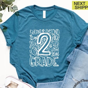 May include: Teal t-shirt with a white graphic that says "Second Grade" with the number 2 in the center. The words "Second" are repeated around the number 2. The shirt is paired with blue jeans and white sneakers.