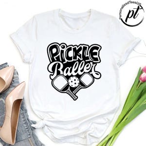 以下が含まれることがあります： 「Pickle Baller」の黒いグラフィックがプリントされた白いTシャツ。デザインには、2つのピックルボールパドルとピックルボールが含まれています。Tシャツはクルーネックで半袖です。