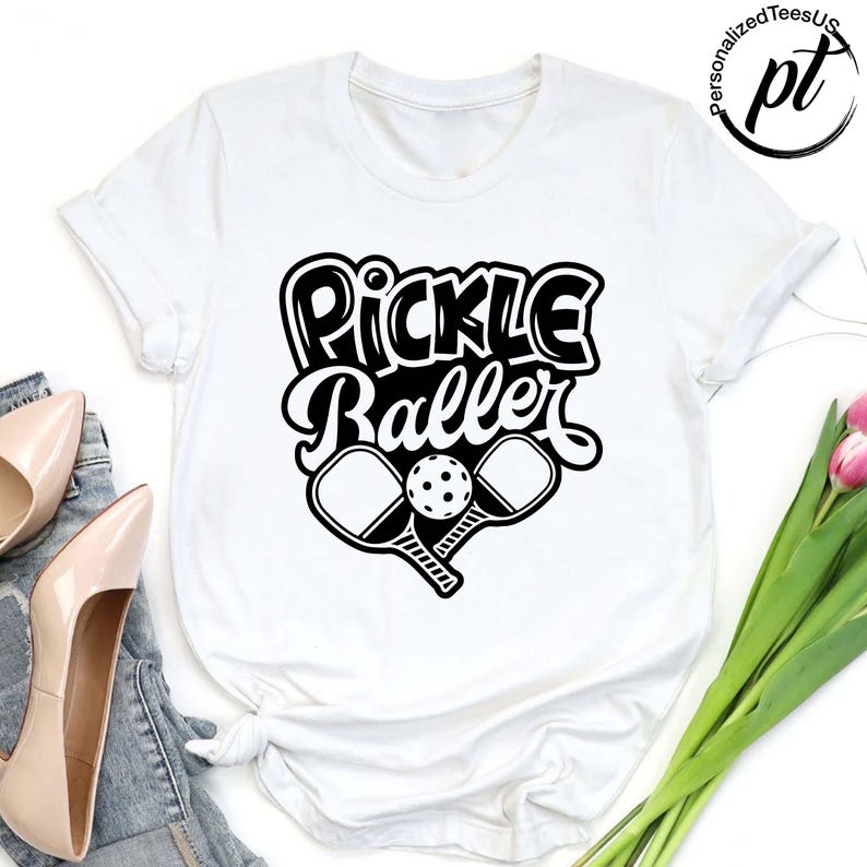 以下が含まれることがあります： 「Pickle Baller」の黒いグラフィックがプリントされた白いTシャツ。デザインには、2つのピックルボールパドルとピックルボールが含まれています。Tシャツはクルーネックで半袖です。
