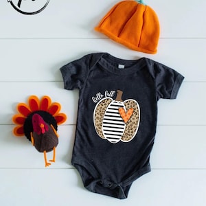 Peut inclure: Un body de b&eacute;b&eacute; gris fonc&eacute; avec un motif de citrouille disant "hello fall". La citrouille a un imprim&eacute; l&eacute;opard, des rayures noires et blanches et un c&oelig;ur orange. Un chapeau citrouille orange et une dinde en feutre sont &eacute;galement dans l'image.