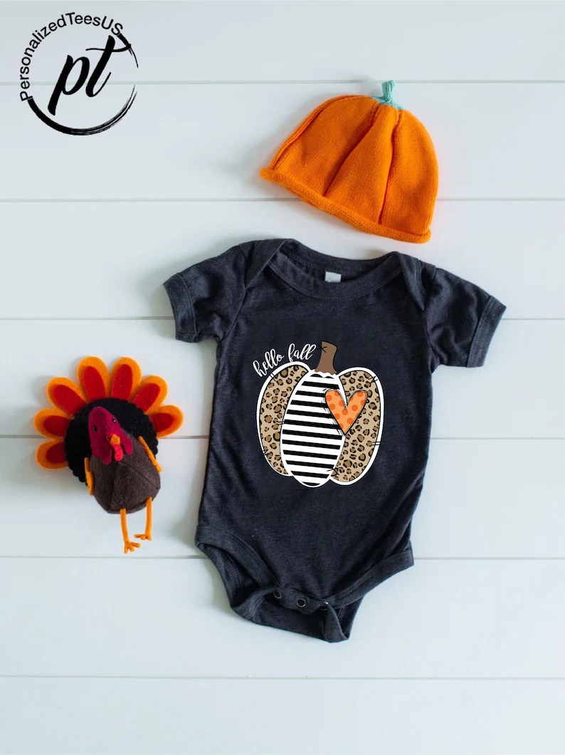 Peut inclure: Un body de b&eacute;b&eacute; gris fonc&eacute; avec un motif de citrouille disant "hello fall". La citrouille a un imprim&eacute; l&eacute;opard, des rayures noires et blanches et un c&oelig;ur orange. Un chapeau citrouille orange et une dinde en feutre sont &eacute;galement dans l'image.