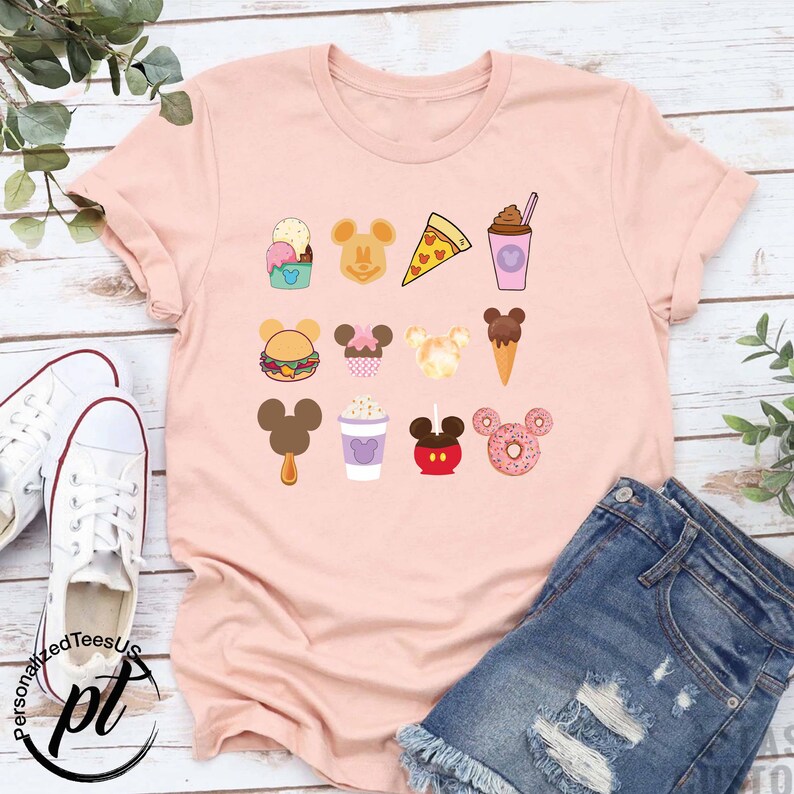 May include: A pink t-shirt with a variety of Disney-themed food and drink icons, including Mickey Mouse ears, ice cream, pizza, and a donut.