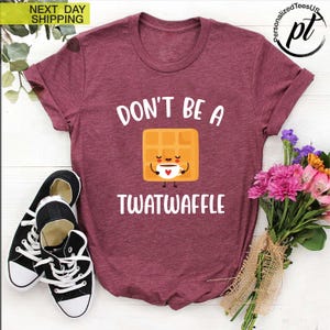 K&ouml;nnte beinhalten: Burgunderfarbenes T-Shirt mit dem wei&szlig;en Text "DON'T BE A TWATWAFFLE". Die Grafik zeigt eine l&auml;chelnde Waffel, die eine Kaffeetasse h&auml;lt. Links sind schwarz-wei&szlig;e Turnschuhe und rechts ein Blumenstrau&szlig;.