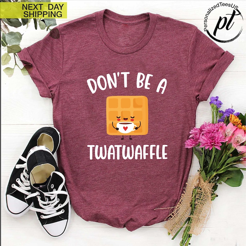K&ouml;nnte beinhalten: Burgunderfarbenes T-Shirt mit dem wei&szlig;en Text "DON'T BE A TWATWAFFLE". Die Grafik zeigt eine l&auml;chelnde Waffel, die eine Kaffeetasse h&auml;lt. Links sind schwarz-wei&szlig;e Turnschuhe und rechts ein Blumenstrau&szlig;.