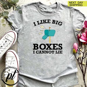 May include: A light gray t-shirt with the text "I LIKE BIG BOXES I CANNOT LIE" in black. The design includes a teal mailbox with a flag and several envelopes. White sneakers are visible on the left.