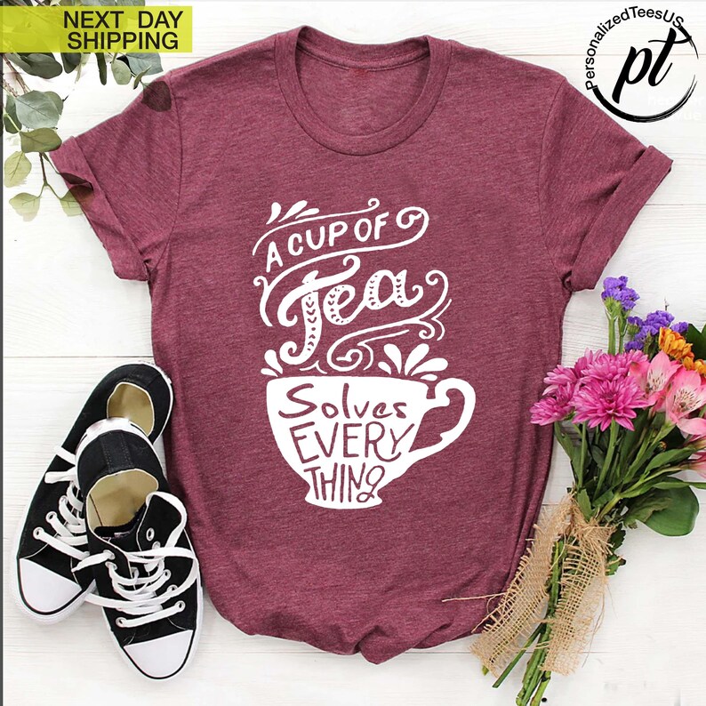 May include: Burgundy t-shirt with the text "A Cup of Tea Solves Everything" in white script. The design features a teacup graphic. Black and white sneakers are placed near the shirt. A bouquet of flowers is on the right.