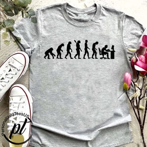 May include: A light gray t-shirt featuring a black graphic depicting the evolution of humans, culminating in a person seated at a computer. The shirt is paired with white canvas sneakers. The text "Personalized TeesUS" and "pt" are visible.
