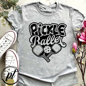 以下が含まれることがあります： 黒いレトロフォントで「Pickle Baller」という文字がプリントされた、ライトグレーのTシャツ。テキストの下には、2つのクロスしたピクルスボールパドルとボールがあります。左側には白いスニーカーが見えます。