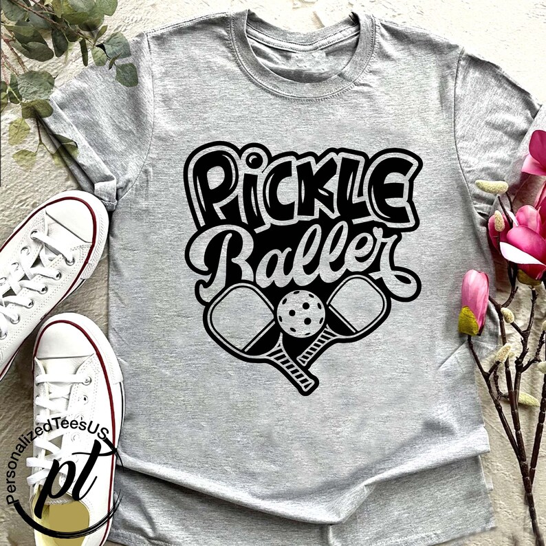 以下が含まれることがあります： 黒いレトロフォントで「Pickle Baller」という文字がプリントされた、ライトグレーのTシャツ。テキストの下には、2つのクロスしたピクルスボールパドルとボールがあります。左側には白いスニーカーが見えます。
