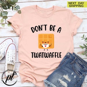 K&ouml;nnte beinhalten: Hellrosa T-Shirt mit dem schwarzen Text "DON'T BE A TWATWAFFLE". Das Design zeigt eine l&auml;chelnde Waffel, die eine Kaffeetasse h&auml;lt. Das Shirt wird mit Jeansshorts und wei&szlig;en Turnschuhen kombiniert.
