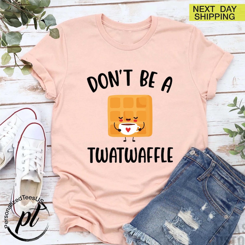 K&ouml;nnte beinhalten: Hellrosa T-Shirt mit dem schwarzen Text "DON'T BE A TWATWAFFLE". Das Design zeigt eine l&auml;chelnde Waffel, die eine Kaffeetasse h&auml;lt. Das Shirt wird mit Jeansshorts und wei&szlig;en Turnschuhen kombiniert.