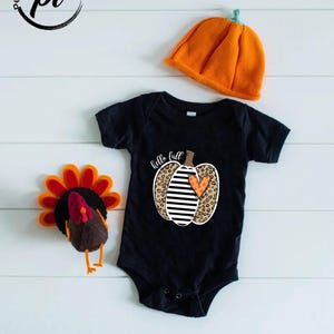 Peut inclure: Un body de b&eacute;b&eacute; noir avec un motif de citrouille qui dit "hello fall". La citrouille a un imprim&eacute; l&eacute;opard, des rayures noires et blanches et un c&oelig;ur orange. Un chapeau citrouille orange et une d&eacute;coration de dinde sont &eacute;galement dans l'image.