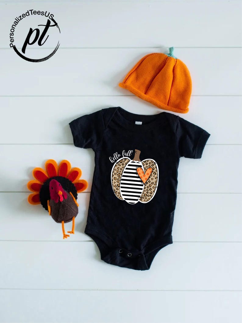 Peut inclure: Un body de b&eacute;b&eacute; noir avec un motif de citrouille qui dit "hello fall". La citrouille a un imprim&eacute; l&eacute;opard, des rayures noires et blanches et un c&oelig;ur orange. Un chapeau citrouille orange et une d&eacute;coration de dinde sont &eacute;galement dans l'image.