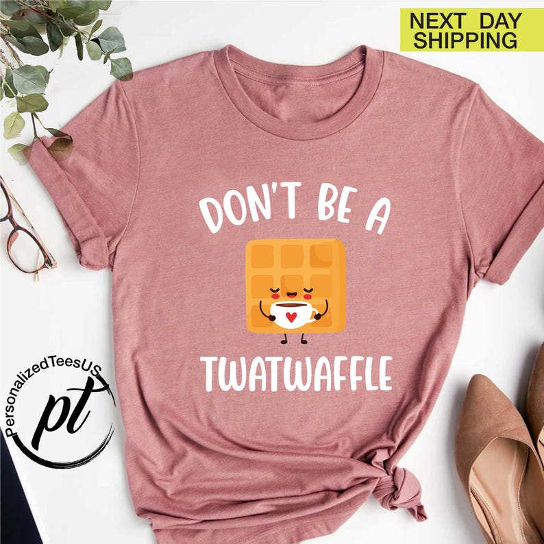 K&ouml;nnte beinhalten: Staubrosa T-Shirt mit dem Text "DON'T BE A TWATWAFFLE" &uuml;ber einem Cartoon-Waffel, der eine Kaffeetasse h&auml;lt. Das Shirt hat kurze &Auml;rmel und einen Rundhalsausschnitt. Das Bild enth&auml;lt auch ein Logo und die Worte "NEXT DAY SHIPPING".