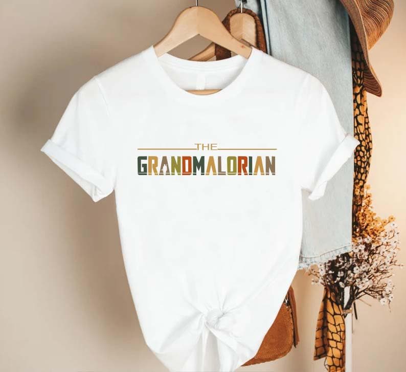 Puede incluir: Camiseta blanca con el texto "THE GRANDMALORIAN" en una fuente colorida de estilo retro. La camiseta tiene mangas cortas y cuello redondo. El dise&ntilde;o presenta una l&iacute;nea horizontal marr&oacute;n encima del texto.